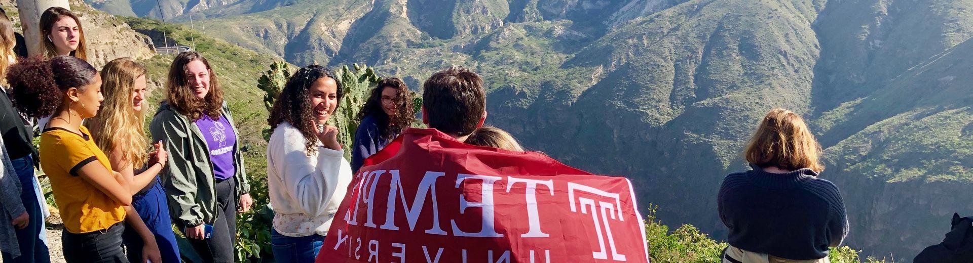 Students on top of a mountain carrying a cherry red Temple flag